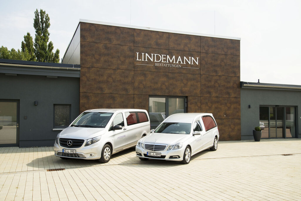 Bestattungsfahrzeug auf gepflastertem Hof vor dem Kolumbarium mit Schriftzug „Lindemann Bestattungen“.