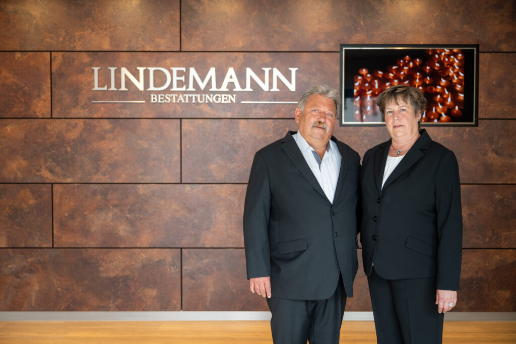 Michael Pflock und Susann Pflock stehen nebeneinander vor der Wand mit dem Schriftzug „Lindemann Bestattungen“.